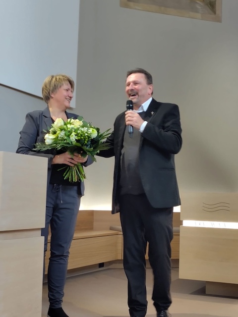 die Innenarchitektin Gudula Be-Pechhold steht mit dem Pfarrer Ludwig Nelles ma Rednerpult und übergibt einen großen Blumenstrauß als Dankeschön
